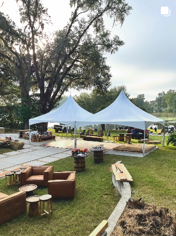 Two high-peak tents with leather lounge furniture and barrel tables at an outdoor party.